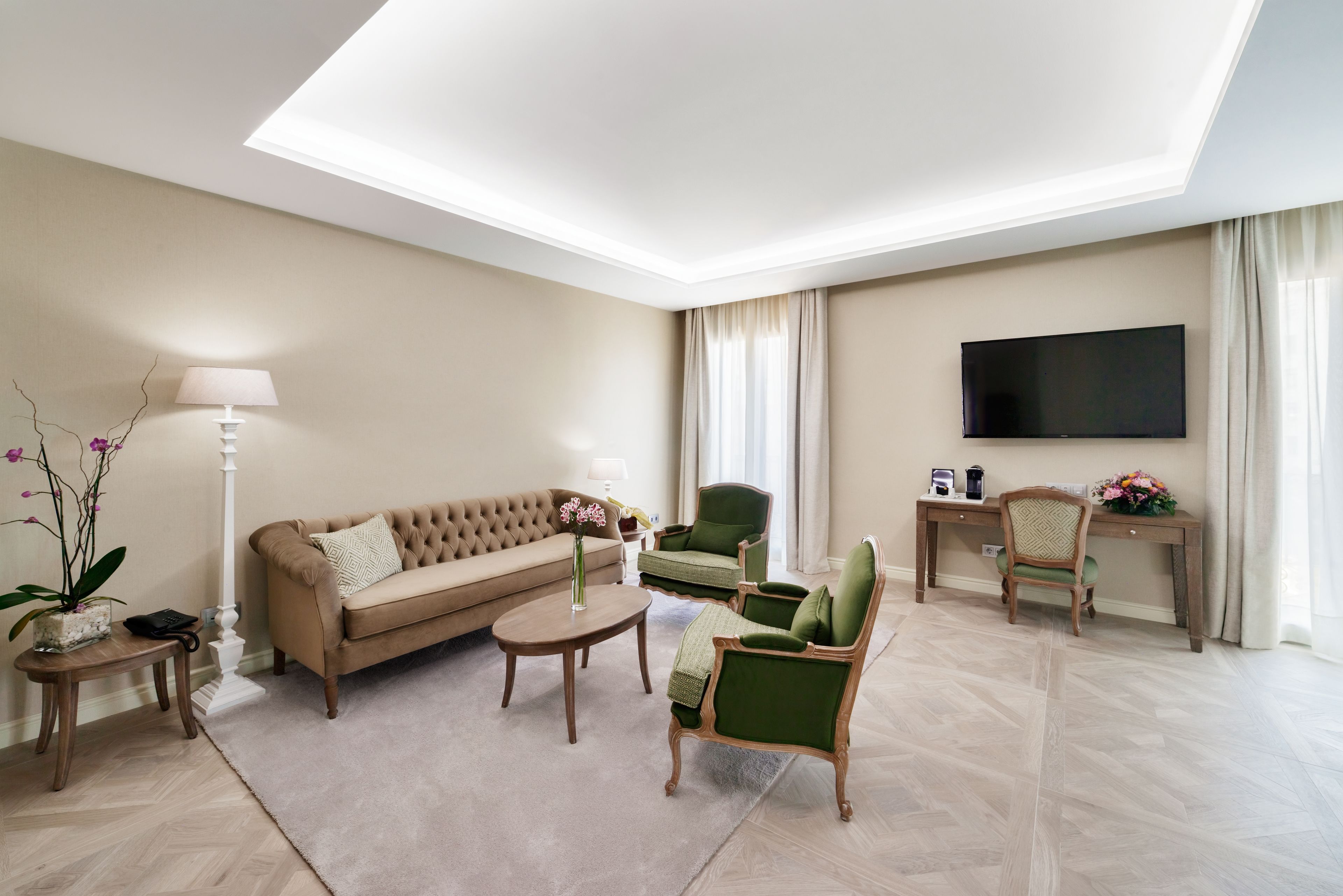 A modern living room with a sofa, armchairs, a coffee table, a TV, and a desk with a lamp and flowers.
