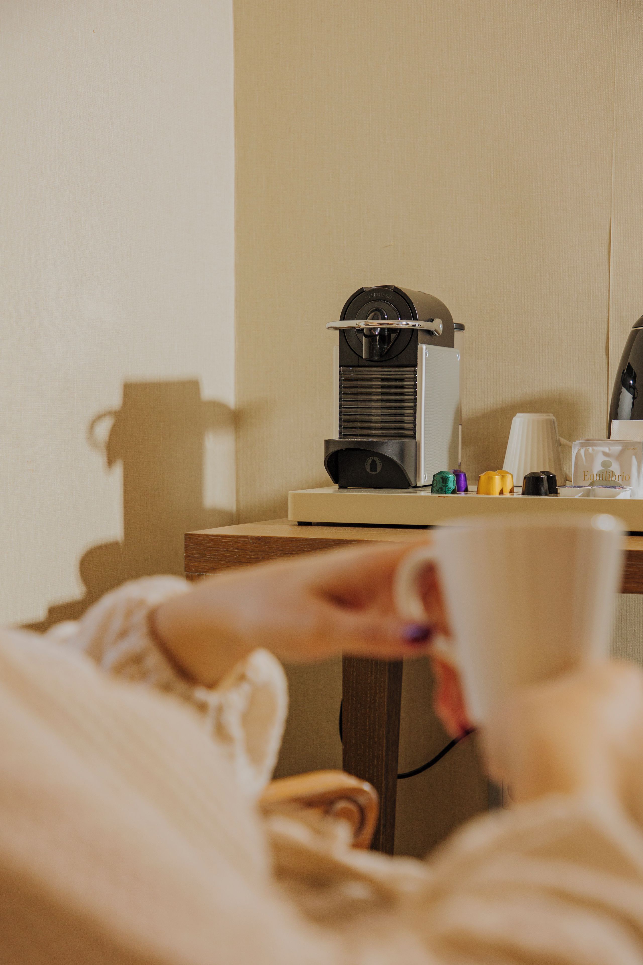 A person holding a cup, a coffee machine, and a small table with various items.