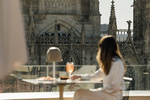 A woman seated at a table with a drink, overlooking a gothic-style building with intricate architecture.