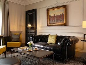 Una habitación con un sofá de cuero negro, un sillón amarillo, una mesa de centro con un florero y dos postres cubiertos con campanas de cristal, y un cuadro de una escena histórica sobre el sofá.
