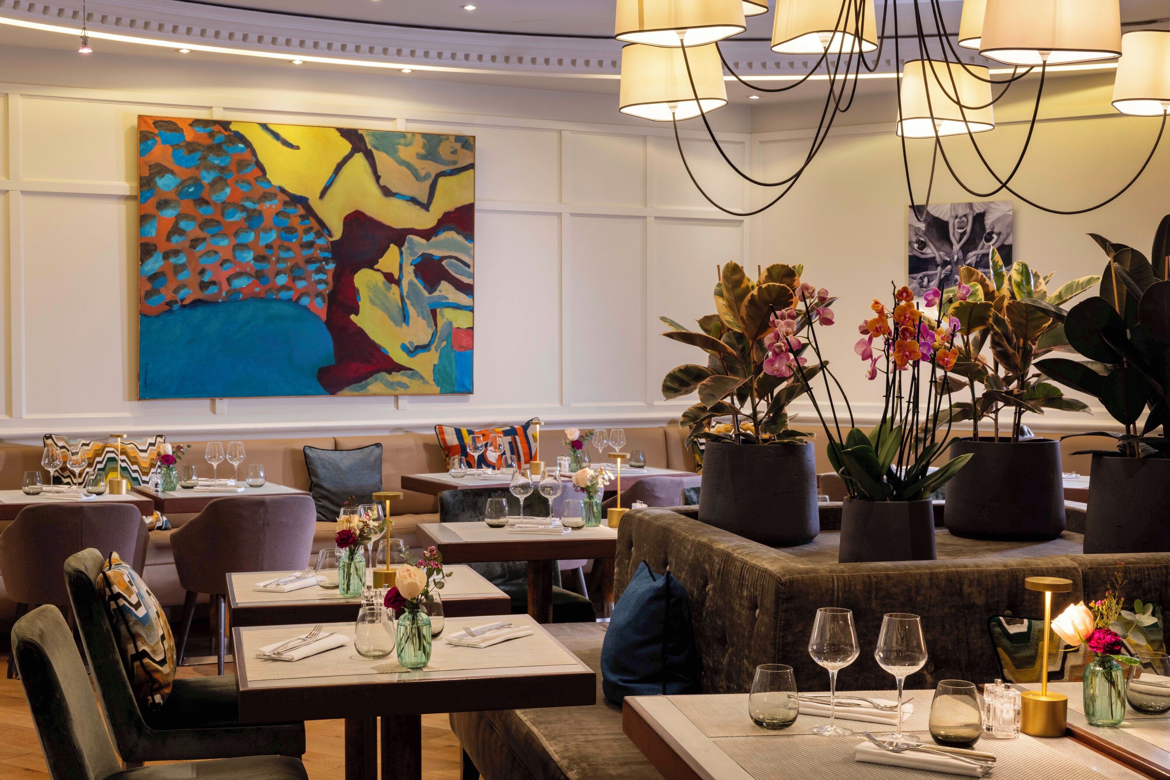 A restaurant interior with tables set for dining, a large abstract painting on the wall, and potted plants.