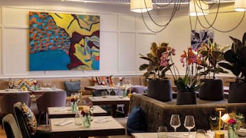 A restaurant interior with tables set for dining, a large abstract painting on the wall, and potted plants.