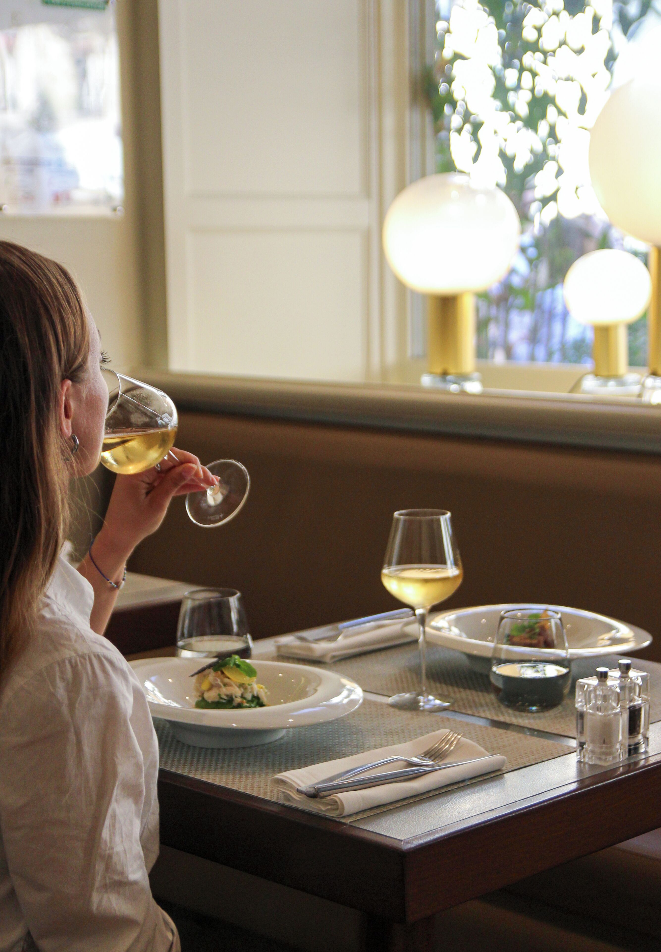 Uma pessoa sentada à mesa com um copo de vinho branco, um prato de comida e uma janela com vista para o verde.