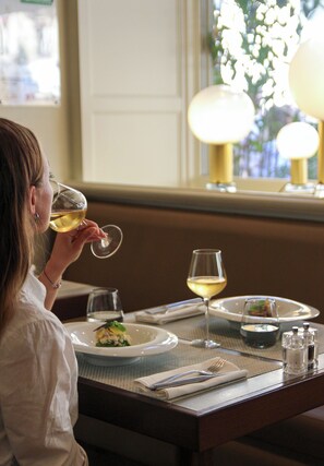 Una persona sentada a una mesa con una copa de vino blanco, un plato de comida y una ventana que da a zonas verdes.