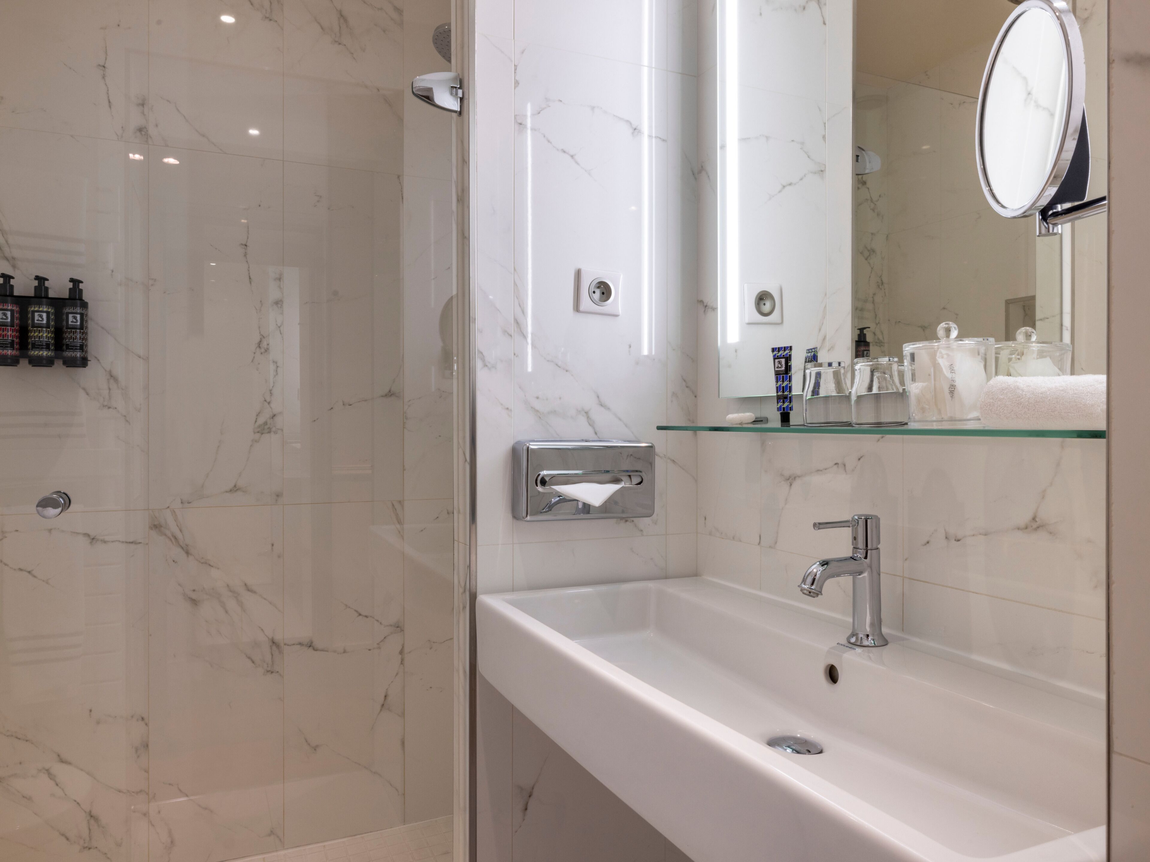 Une salle de bain avec un mur en marbre, une étagère en verre où sont rangés les articles de toilette, un lavabo blanc et un miroir.