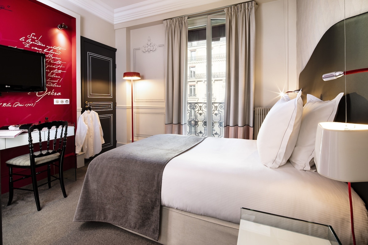 Una habitación de hotel con cama, escritorio, silla, televisor y ventana con cortinas.
