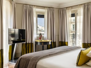 Una habitación de hotel con una cama, un televisor, una mesa redonda con un florero y cortinas.