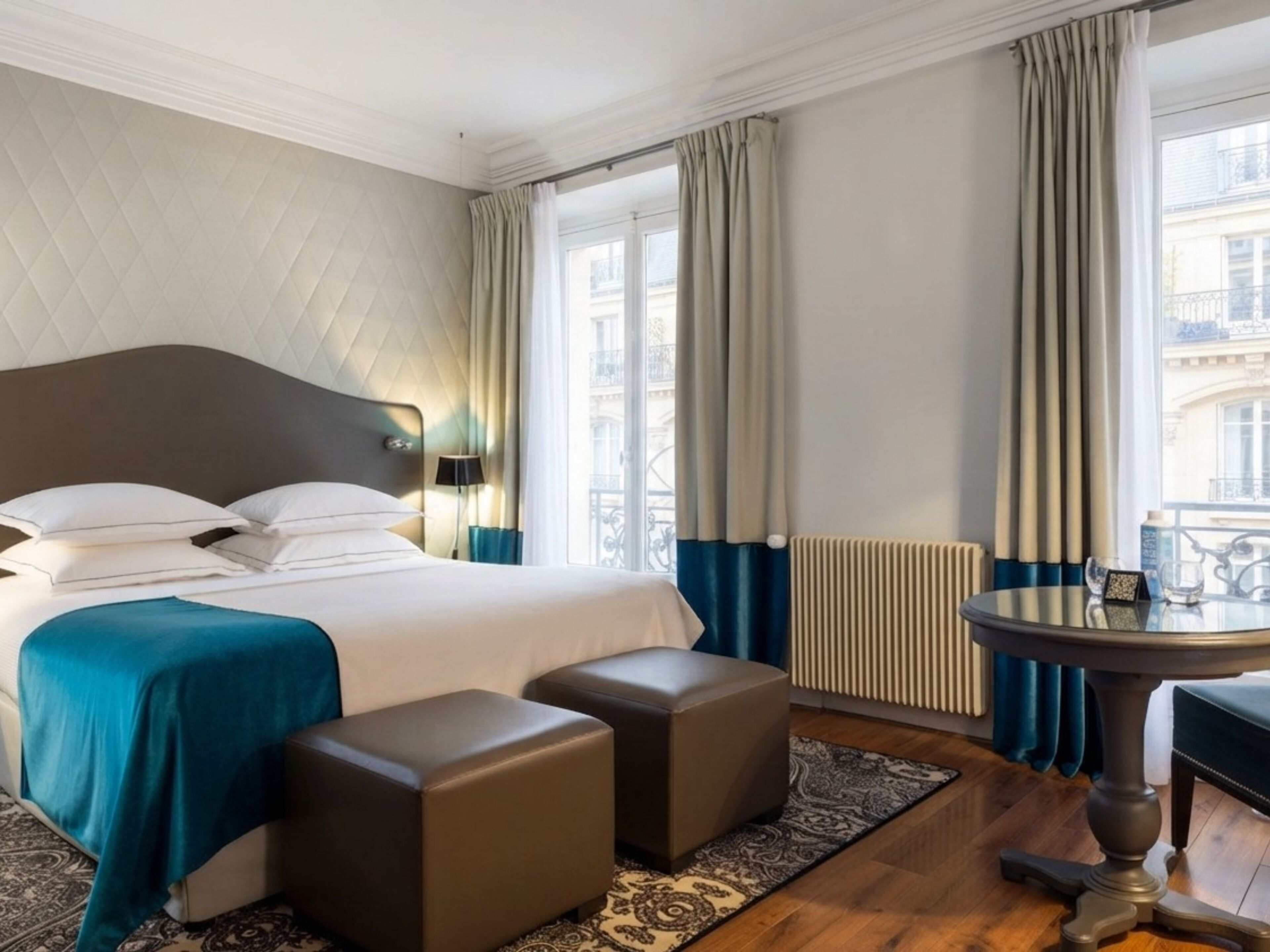Une chambre d’hôtel avec un lit, deux poufs, une table et une vue sur la ville.