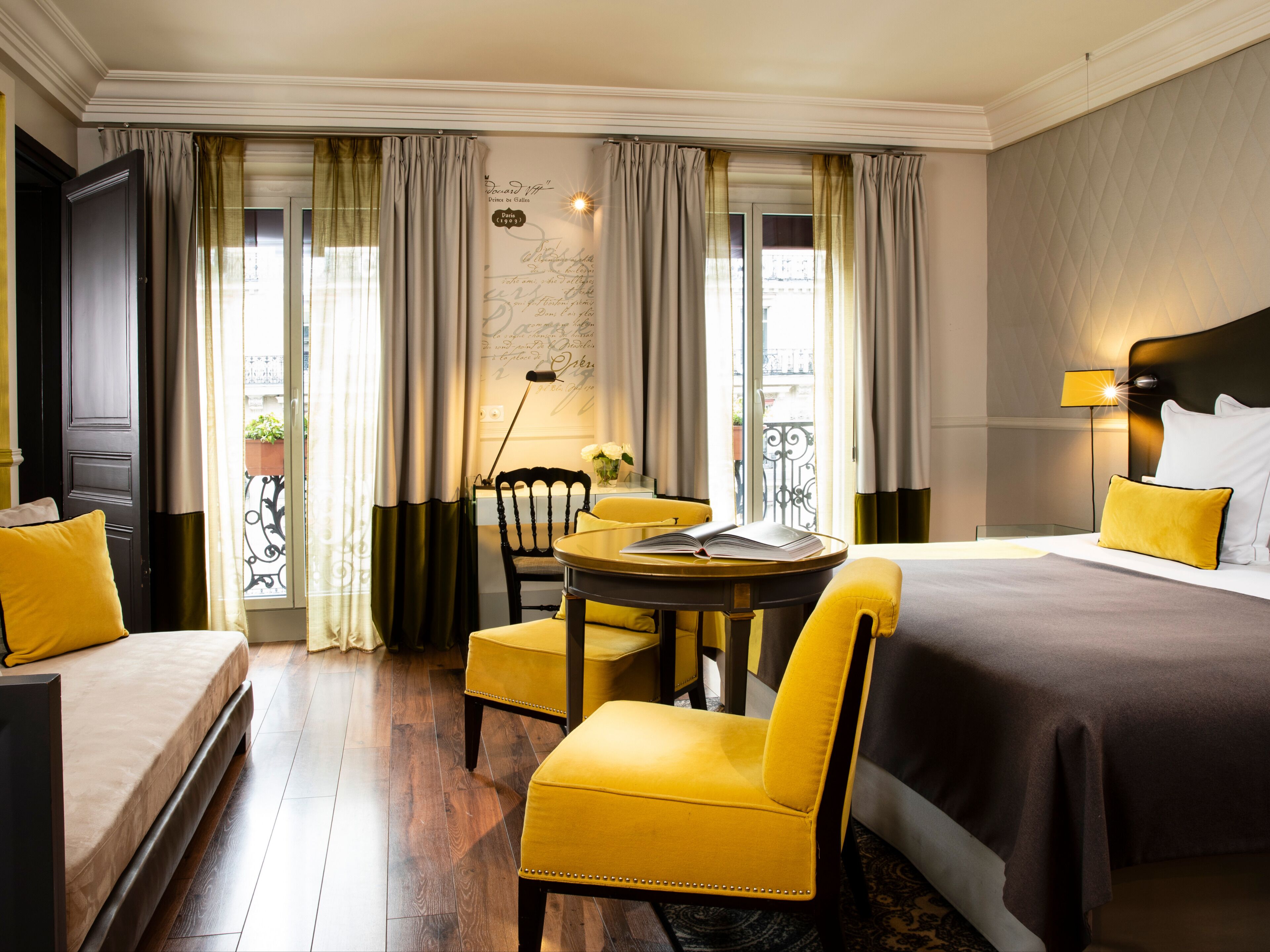 A hotel room with a bed, two yellow armchairs, a small table with a book, a nightstand with a lamp, and a view of the outside balcony.