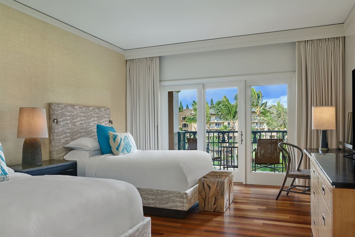 A hotel room with two beds, a desk, and a view of a balcony with palm trees.