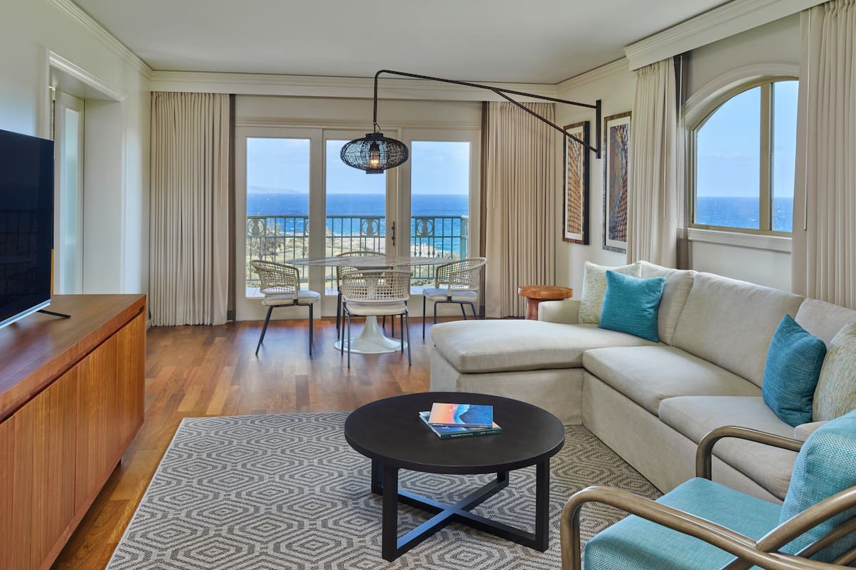 A spacious living room with a sofa, a coffee table, and a TV on a wooden cabinet. There is a balcony with a view of the ocean.
