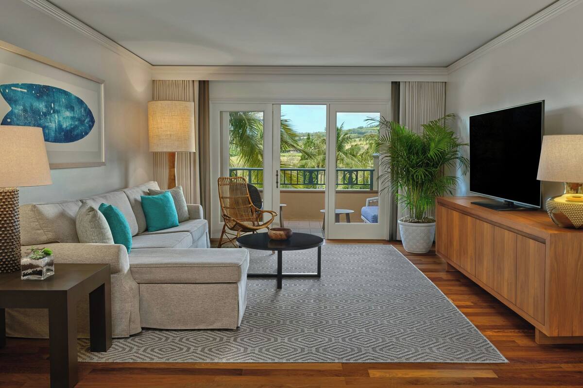 A modern living room with a sofa, a coffee table, a TV, and a balcony with a view of palm trees.