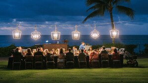 Un área de comedor al aire libre con mesas y sillas, palmeras y luces colgantes, con vista al océano y al cielo.