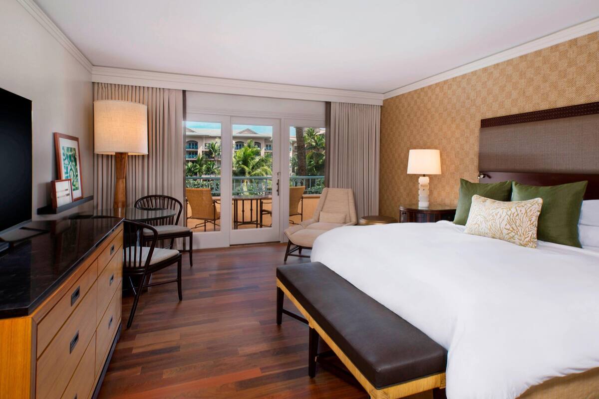 A hotel room with a large bed, a desk with a TV, a chair, a bench, and a balcony with a view of palm trees.