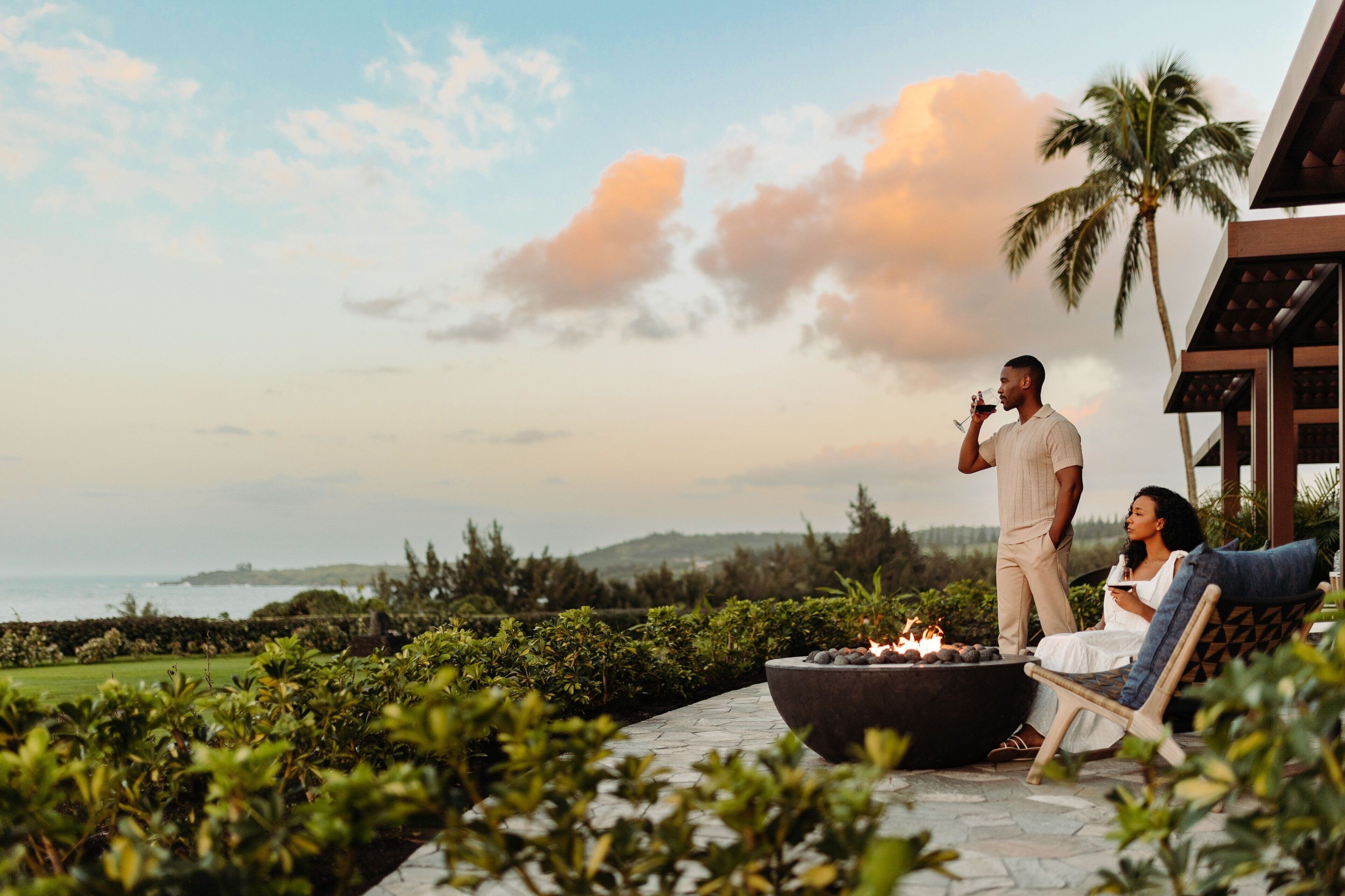 A couple relaxing by a fire pit with a view of the ocean and palm trees.