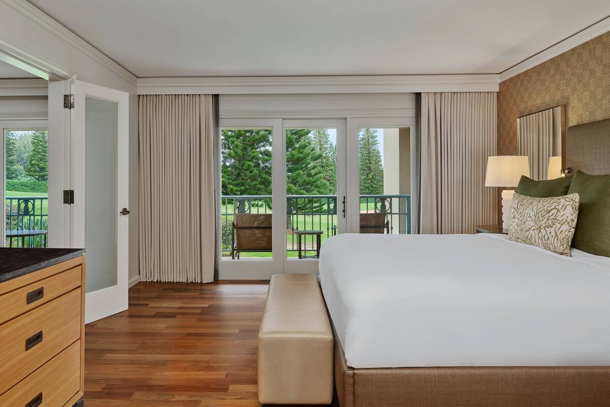 A hotel room with a large bed, a bench, a dresser, and a balcony with a view of trees.