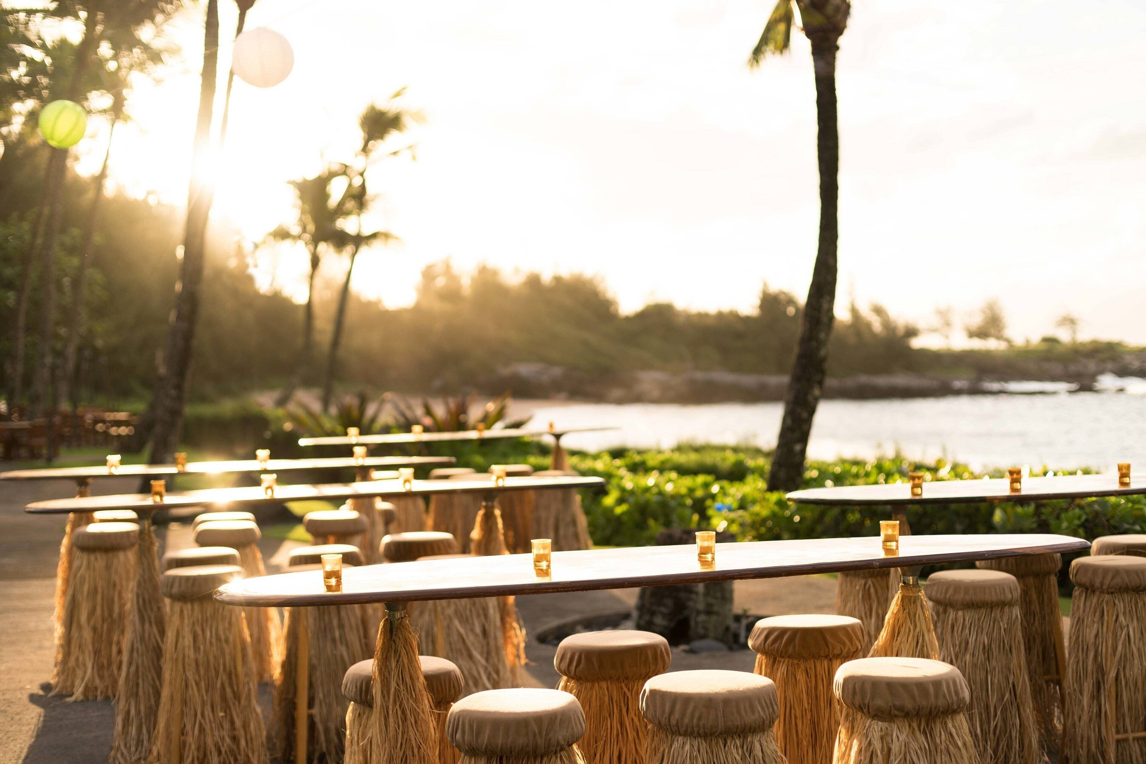 Outdoor tables with stools, arranged in rows, overlooking a body of water with palm trees in the background.