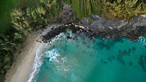 Vista aérea de una zona costera con playa arenosa, aguas turquesas cristalinas y densa vegetación verde.