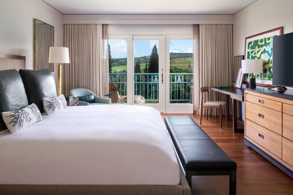A hotel room with a large bed, a desk, a chair, a TV, and a balcony with a scenic view.
