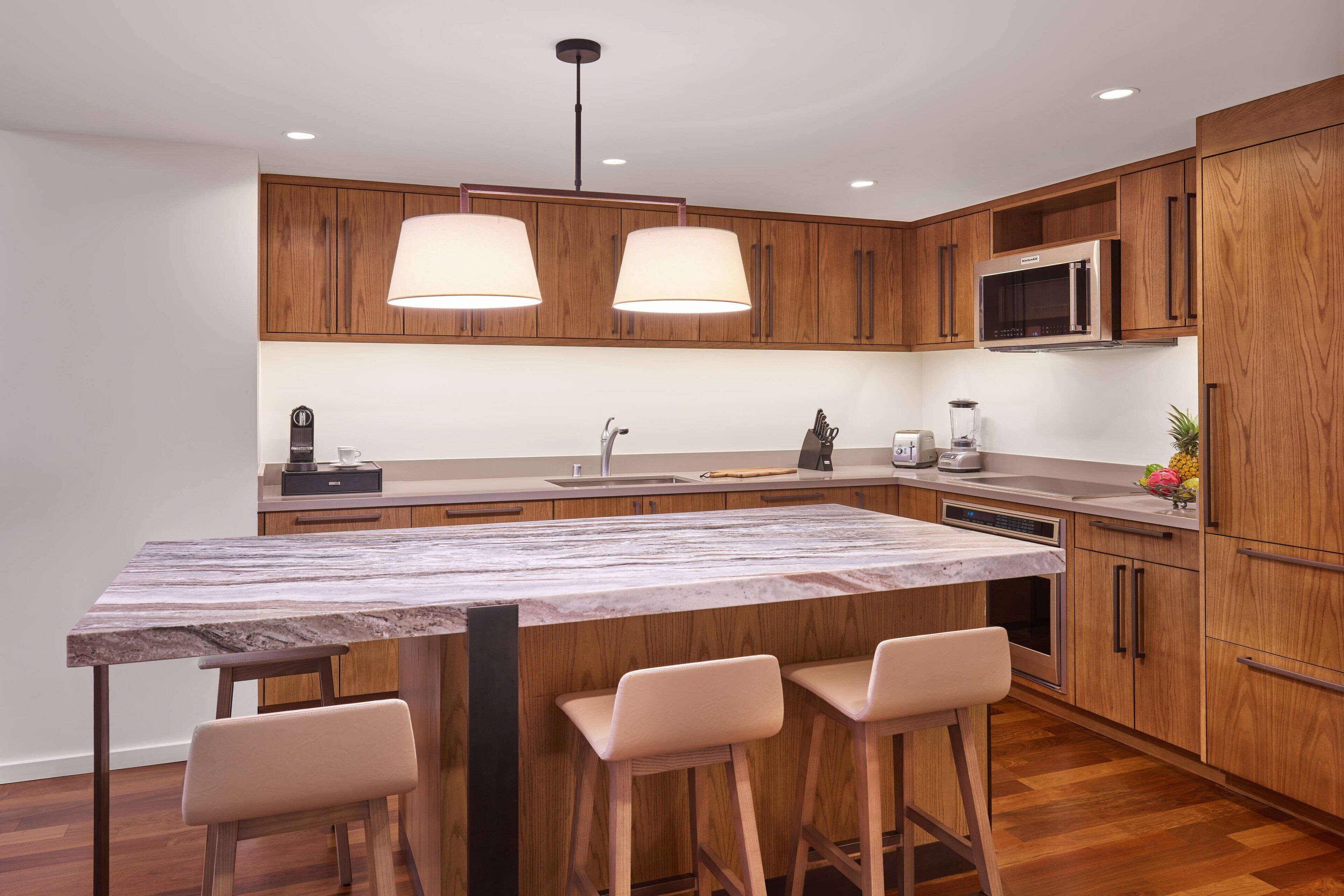 A modern kitchen with wooden cabinets, a marble countertop, a built-in microwave, and a large island with bar stools.