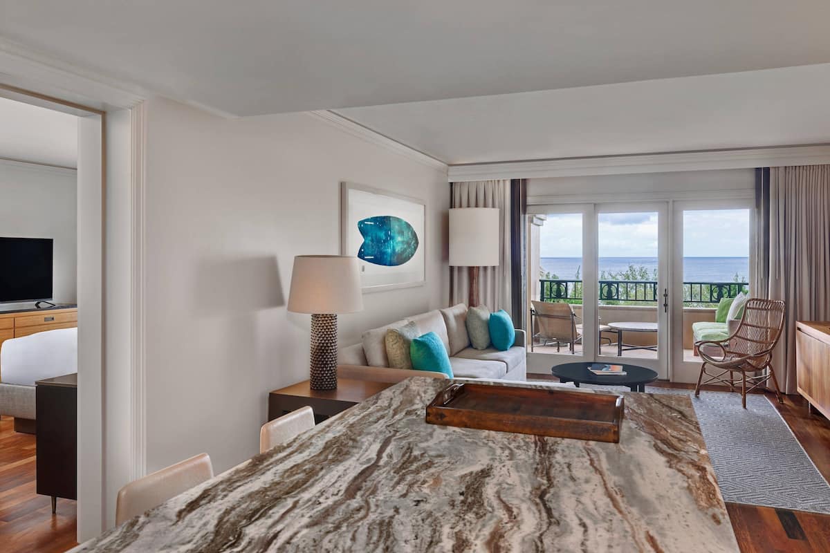A hotel room with a marble dining table, a sofa with colorful cushions, a television, and a balcony with a view of the sea.