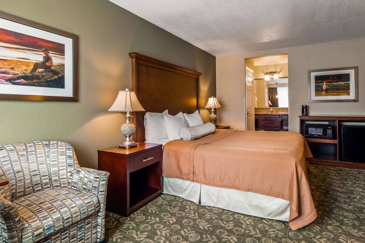 A hotel room with a bed, a chair, a desk, a lamp, a television, and a painting on the wall.