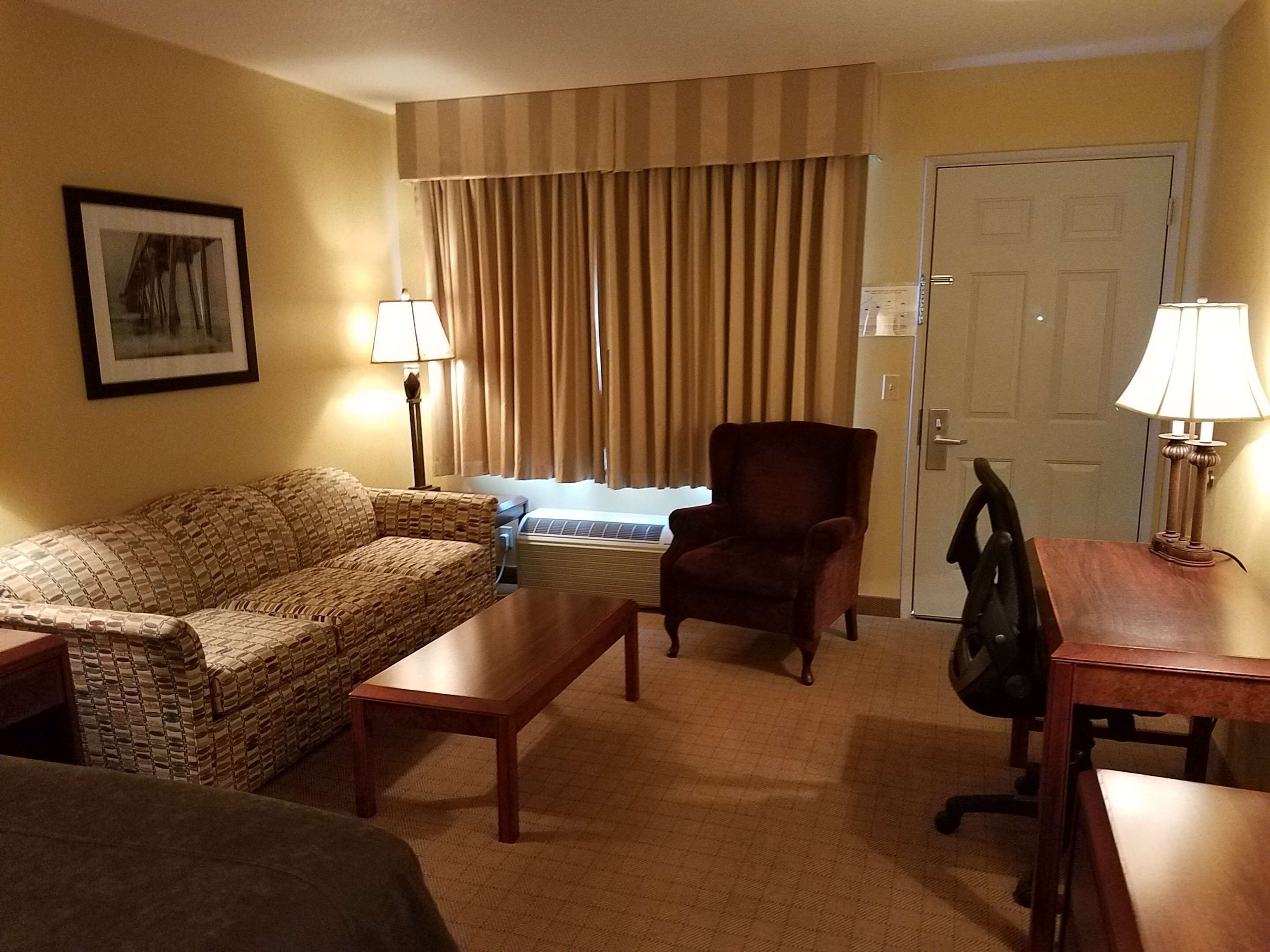 A hotel room with a patterned sofa, a wooden armchair, a small table, and a desk with a lamp. There is a window with curtains and a door in the background.