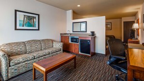 A compact hotel room with a sofa, a small wooden table, a kitchenette with a microwave and mini-fridge, a desk with a lamp, and a framed picture on the wall.