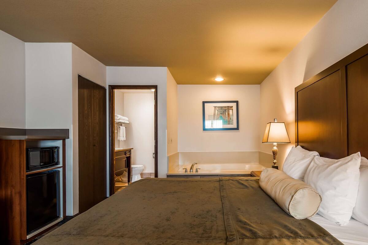 A hotel room with a bed, a wooden headboard, a bedside lamp, a bathroom with a sink and toilet, and a microwave in the corner.