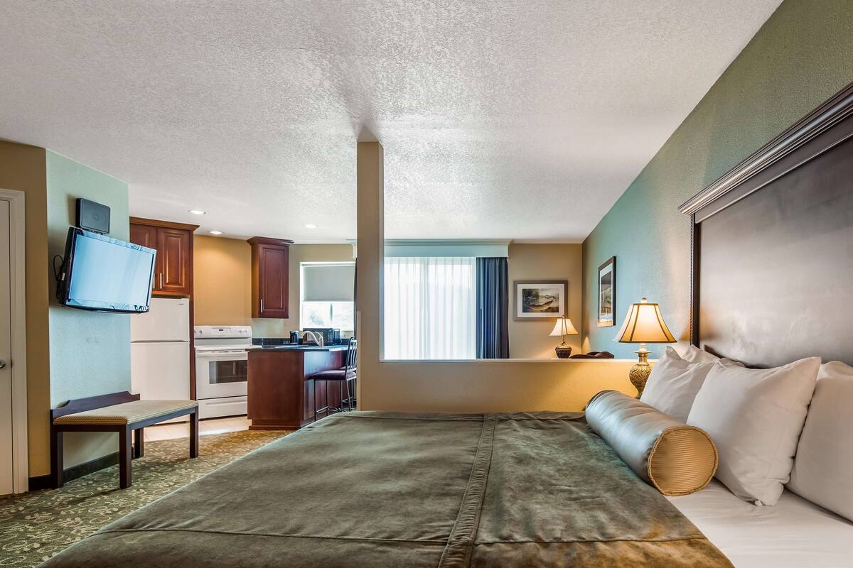 A hotel room with a bed, a desk, a kitchenette, and a television.