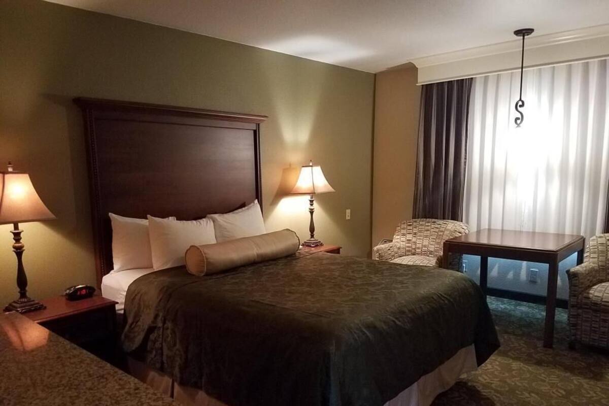 A hotel room with a large bed, two bedside tables with lamps, a chair, a small table, and a view of the city through the window.