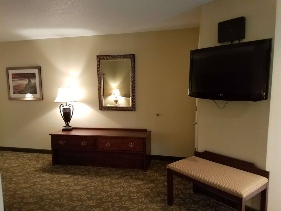 A hotel room with a wooden bench, a TV mounted on the wall, a mirror, a lamp, and a framed picture on the wall.