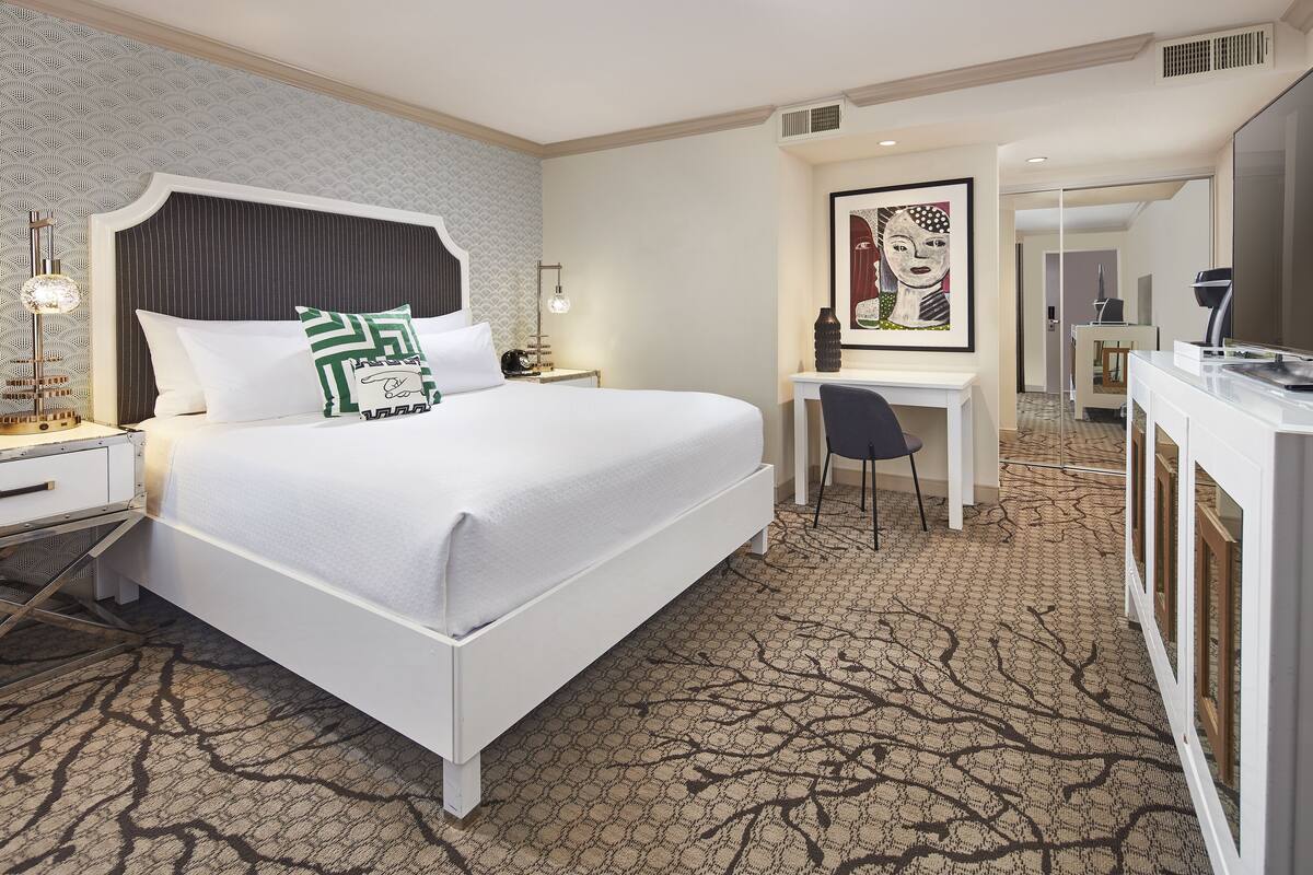 A hotel room with a large bed, a desk, and a chair. There is a painting on the wall and a lamp on the nightstand.