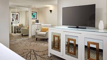 A hotel room with a large flat-screen TV, a glass-top coffee table, a white cabinet with mirrored doors, a patterned carpet, and a chair with a yellow pillow.