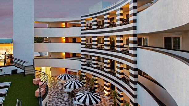 Un hotel de varios niveles con fachada curva, zona de estar al aire libre con sombrillas y un piso con diseño de cuadros.