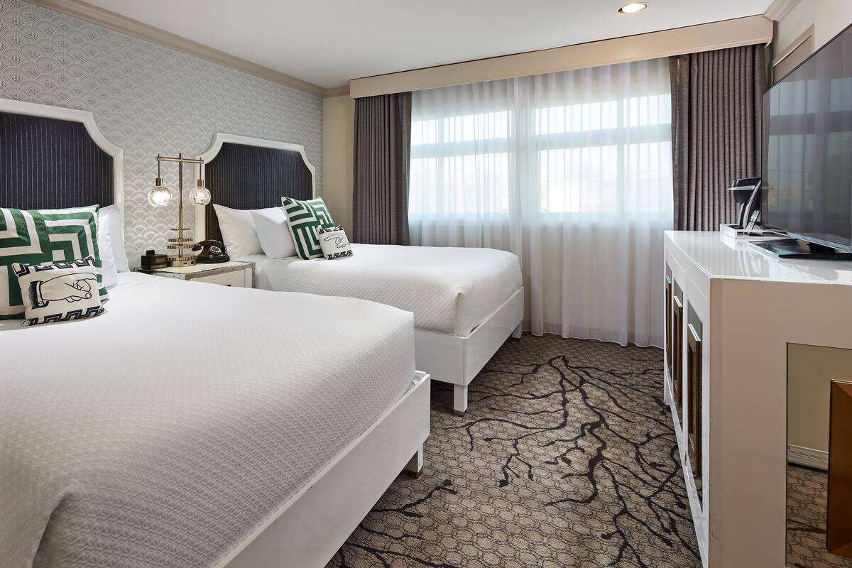 A hotel room with two beds, a desk, a TV, and a large window with curtains.