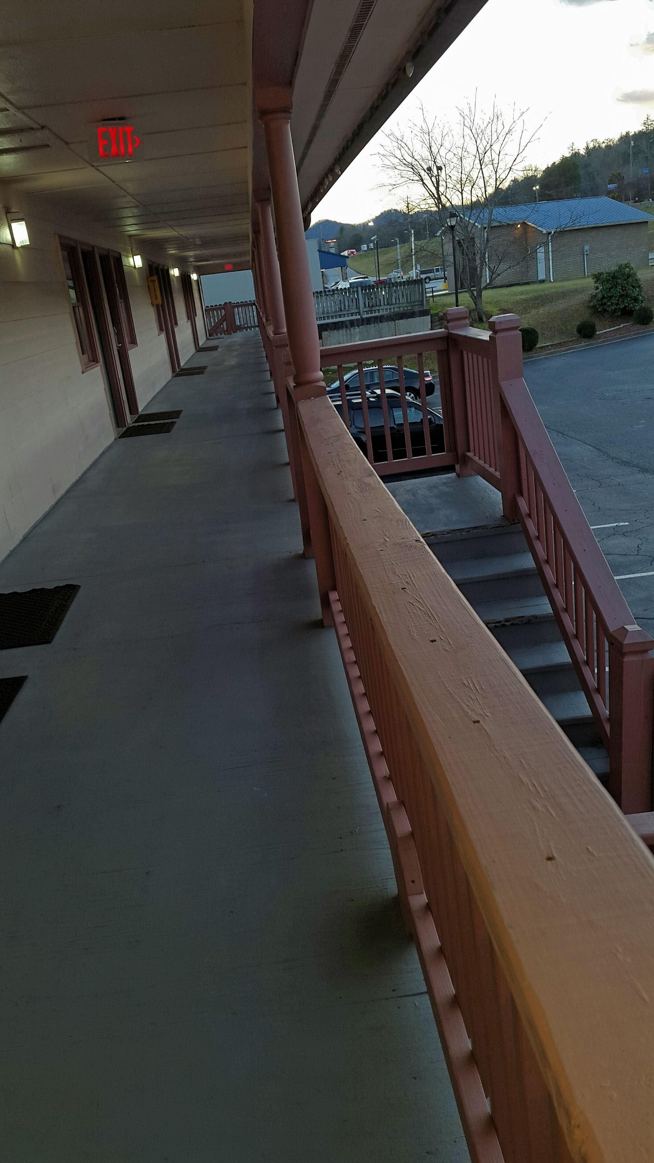 Een overdekte doorgang met een rode reling, een vluchtwegbord en een uitzicht op een parkeerplaats en gebouwen in de verte.