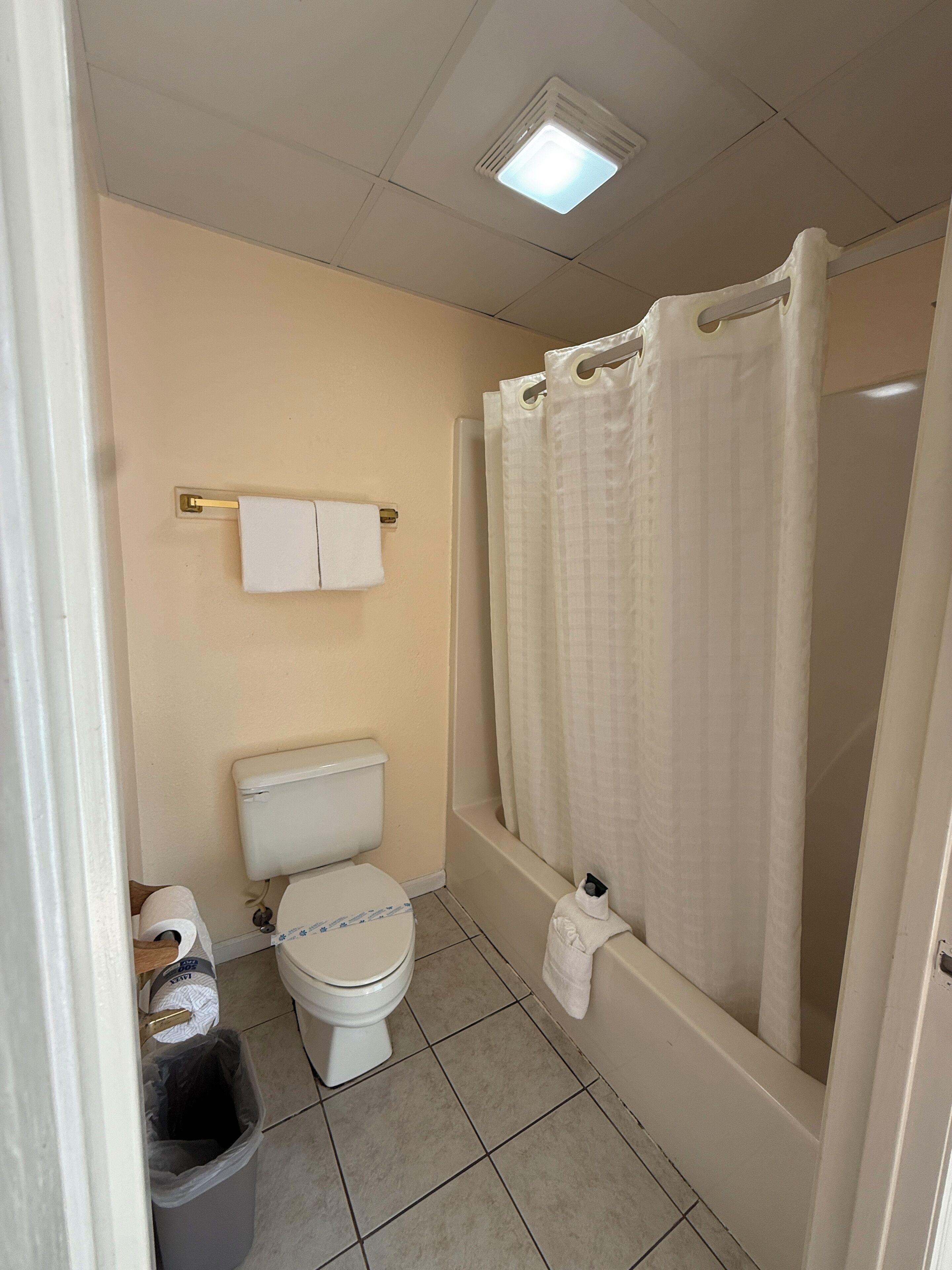 A bathroom with a toilet, a bathtub with a shower curtain, a towel rack, and a trash bin.