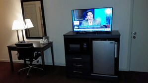 Una habitación de hotel con escritorio, televisor mostrando un noticiero, microondas y refrigerador.