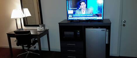 Una habitación de hotel con escritorio, televisor mostrando un noticiero, microondas y refrigerador.