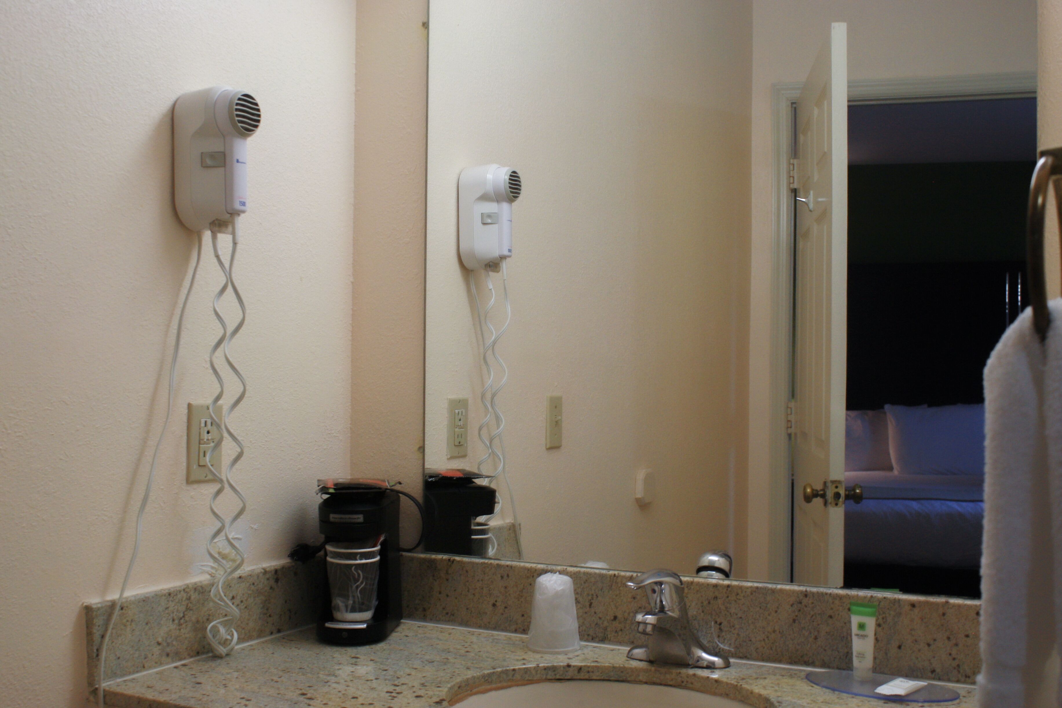 A hotel bathroom with a sink, two hair dryers mounted on the wall, a coffee maker, and a mirror reflecting a bed and a door.
