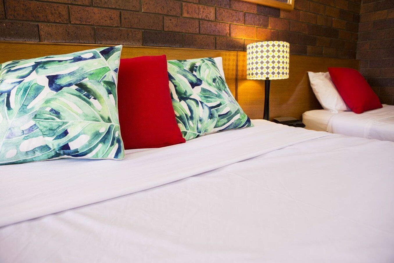 A hotel room with a brick wall, a bed with white bedding, red and green patterned pillows, and a lamp with a patterned shade.