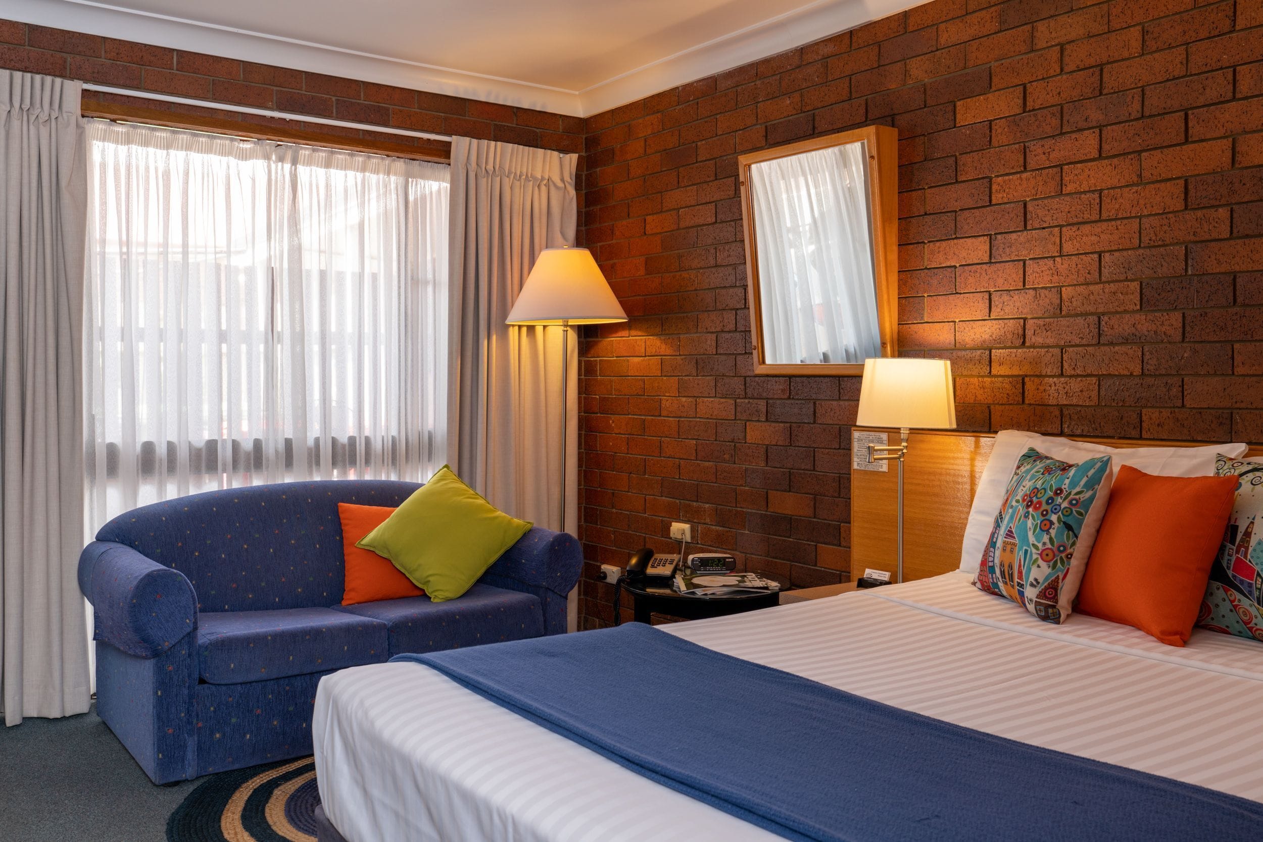 A hotel room with a brick wall, a blue sofa with a green pillow, a bed with patterned pillows, and a nightstand with a lamp.