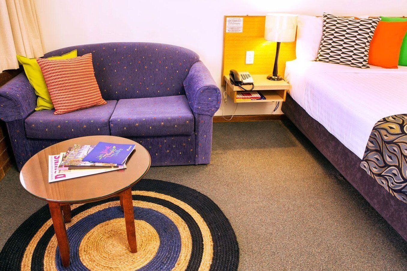 A hotel room with a purple sofa, a small table with magazines, a lamp, and a bed with patterned pillows.