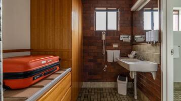 Un baño pequeño con una maleta roja en un estante, un lavamanos, un espejo y una ventana con una toalla colgada en la pared.