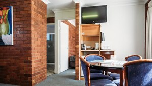 Habitación de hotel con pared de ladrillo, televisor de pantalla plana sobre un escritorio de madera y zona de comedor con sillas tapizadas en azul.