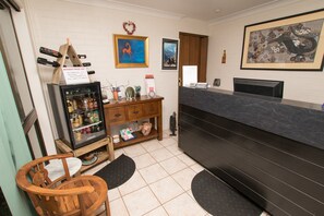 A reception area with a wooden desk, a refrigerator stocked with beverages, a small shelf with decorative items, and a framed picture on the wall.