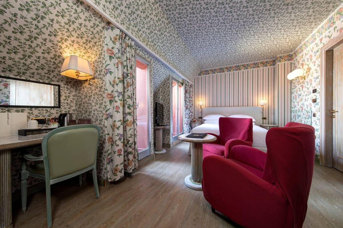 A hotel room with floral wallpaper, a bed, a desk with a chair, and a red armchair.