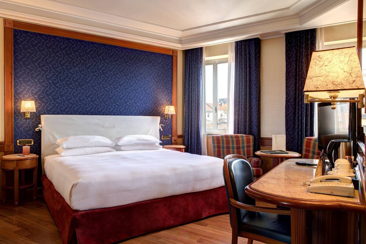 A hotel room with a large bed, a wooden desk, a chair, a television, and a window with curtains.