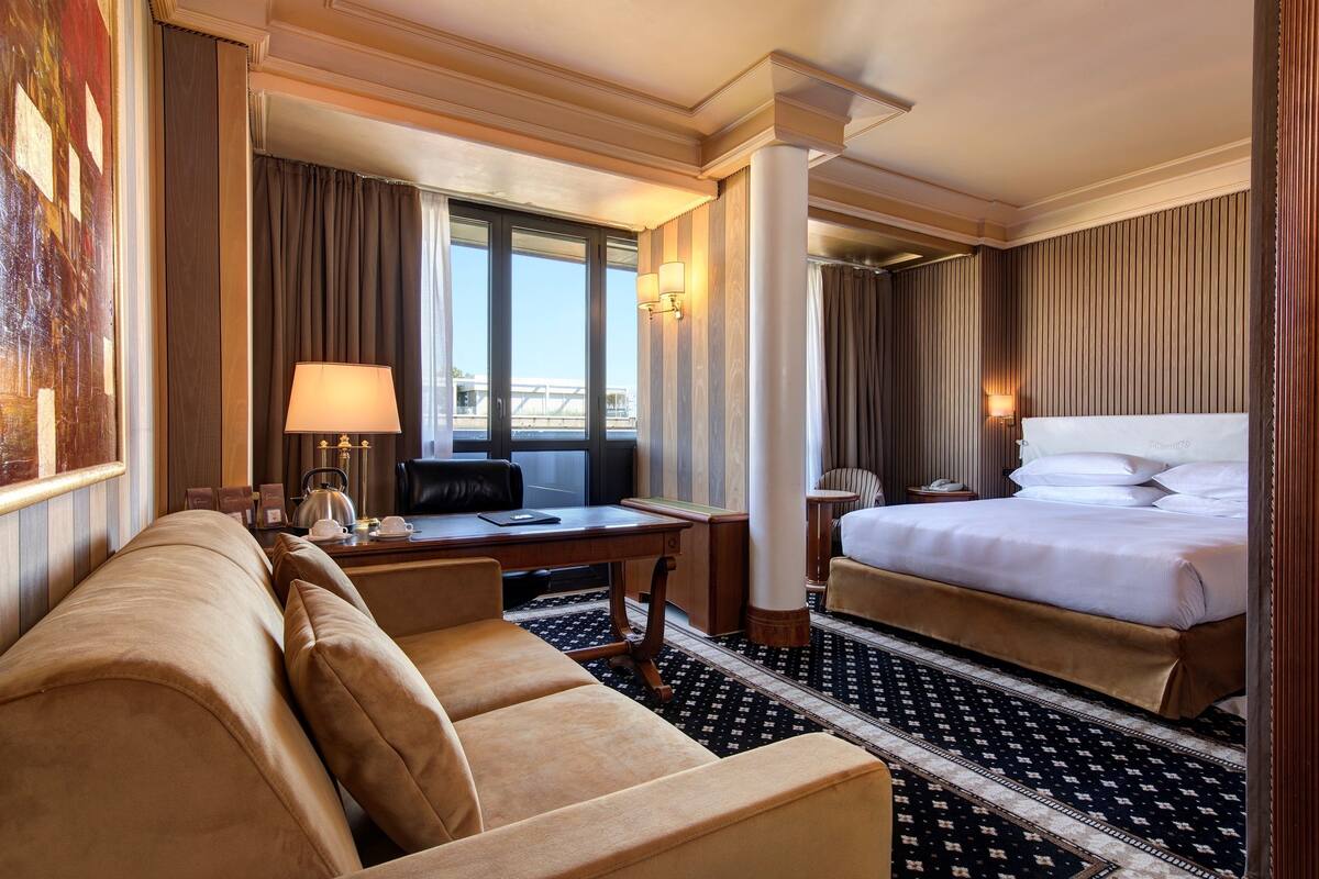 A hotel room with a sofa, a desk with a lamp, a bed, and a view of the city.